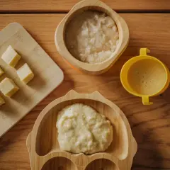Pierwsza kaszka dla niemowlaka karmionego piersią. Na drewnianym blacie leżą kosteczki banana, bambusowa miseczka z kaszką i żółty kubeczek.