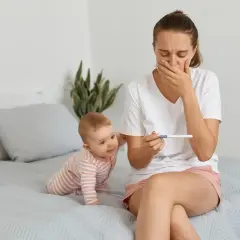 Zaskoczona mama z niemowlakiem na rękach trzyma test ciążowy. Zastanawia się, po jakim czasie można zajść w ciążę po porodzie naturalnym.