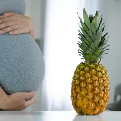 Ciężarna kobieta w szarej sukience trzyma się za brzuch obok ananasa.