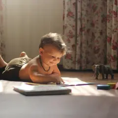 Małe dziecko leżące na brzuchu, podnoszące główkę, z zaciekawieniem przegląda książeczkę. Obok leżą zabawki: pociąg, słoń i wieża z kółek.