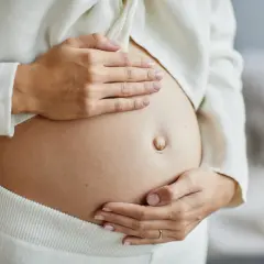 Zbliżenie na ciążowy brzuch kobiety, która delikatnie go obejmuje dłońmi.