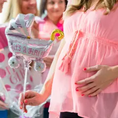 Ciężarna kobieta w różowej sukience trzyma balon w kształcie wózka z napisem "it's a girl!".