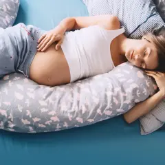Kobieta w ciąży leży na brzuchu, otulona poduszką. Jej dłoń delikatnie spoczywa na zaokrąglonym brzuchu.