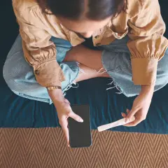 Kobieta siedzi ze skrzyżowanymi nogami, trzymając w dłoniach telefon i test ciążowy.