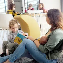 Mama karmi piersią niemowlę, podczas gdy starsze dziecko czyta książkę. To piękny przykład karmienia mieszanego w rodzinnej atmosferze.