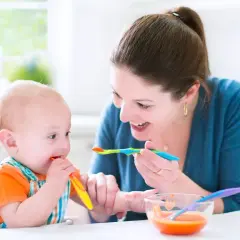 Mama karmi niemowlę, które próbuje samodzielnie jeść. To obrazek z tabeli żywienia 7-miesięcznego dziecka.