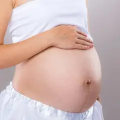 Brzuch kobiety w białej bluzce i spódnicy. Dłoń spoczywa na brzuchu, sugerując który tydzień ciąży.