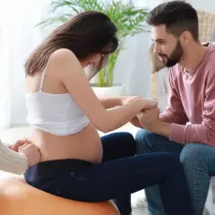 Kobieta w ciąży na piłce, czuć ruchy dziecka. Partner i doula wspierają ją.