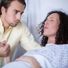 Mężczyzna trzyma za rękę kobietę w szpitalnym łóżku, która przeżywa skurcze. To już 40 tydzień ciąży, a poród jest blisko.