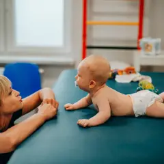Fizjoterapeutka zachęca dziecko do obrotów na boki. Maluch leży na brzuchu, gotowy do nauki nowych umiejętności.