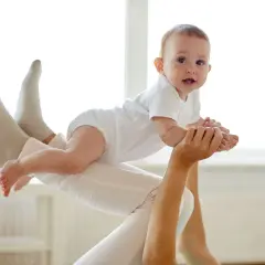 Radość z zabaw z półrocznym dzieckiem. Maluch w białym body unosi się w powietrzu, trzymany przez rodzica.
