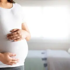 Kobieta w białej koszulce trzyma dłonie na zaokrąglonym brzuchu ciążowym.