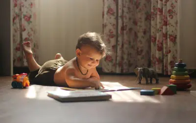Małe dziecko leżące na brzuchu, podnoszące główkę, z zaciekawieniem przegląda książeczkę. Obok leżą zabawki: pociąg, słoń i wieża z kółek.