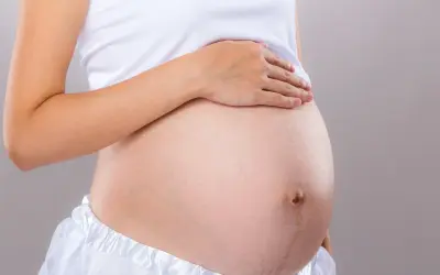 Brzuch kobiety w białej bluzce i spódnicy. Dłoń spoczywa na brzuchu, sugerując który tydzień ciąży.