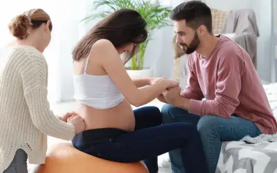 Kobieta w ciąży na piłce, czuć ruchy dziecka. Partner i doula wspierają ją.