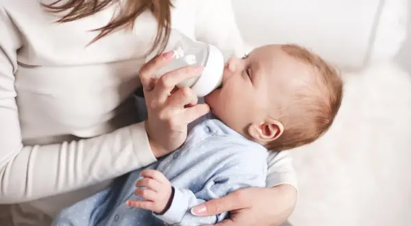 Delikatne karmienie dziecka butelką. Maluch w niebieskim pajacyku pije mleko, czując się bezpiecznie w ramionach mamy.