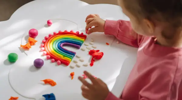 Dziecko tworzy tęczę z plasteliny, rozwijając kreatywne zabawy dla dzieci w domu. Kolorowe kuleczki i paski plasteliny tworzą magiczny świat.