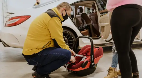 Mężczyzna w żółtej kurtce zapina pasy w czerwonej wkładce do nosidełka Maxi-Cosi, przygotowując ją do podróży samochodem.