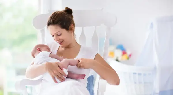Uśmiechnięta mama kołysze noworodka. W tle widać łóżeczko i kwiaty. To może być moment, gdy zastanawiasz się, jak zatrzymać laktację.