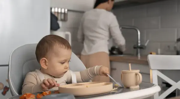 Dziecko siedzi w krzesełku do karmienia, próbując samodzielnie jeść. W tle mama przygotowuje posiłek.
