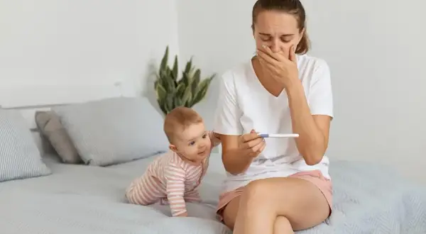 Zaskoczona mama z niemowlakiem na rękach trzyma test ciążowy. Zastanawia się, po jakim czasie można zajść w ciążę po porodzie naturalnym.