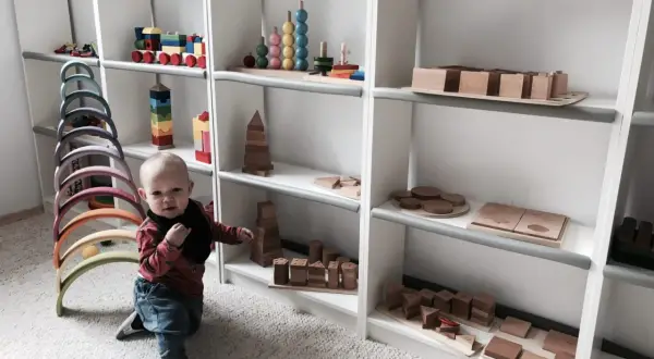 Małe dziecko w bawialni Montessori, otoczone drewnianymi zabawkami edukacyjnymi, takimi jak tęczowe łuki i klocki.