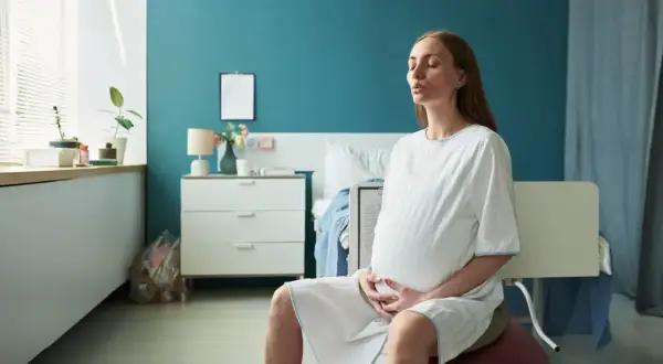 Kobieta w ciąży na piłce do ćwiczeń, z zamkniętymi oczami, oddycha głęboko. Czy twardnienie brzucha to skurcze? Pokój porodowy.