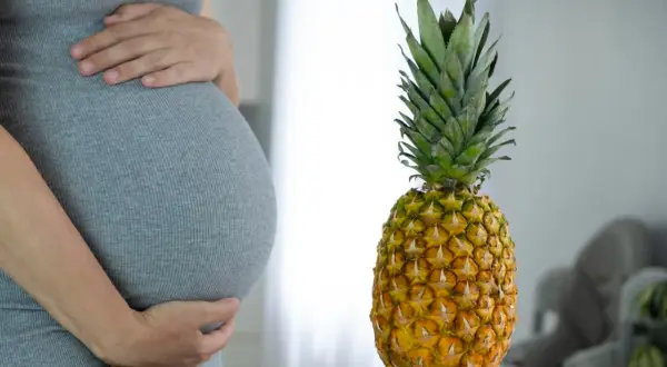 Ciężarna kobieta w szarej sukience trzyma się za brzuch obok ananasa.
