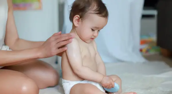 Mama smaruje kremem ramię dziecka, które siedzi w prawidłowym siadzie. Dziecko trzyma w rączce niebieską kostkę.