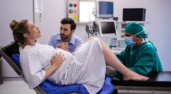 Kobieta rodzi, wspierana przez partnera i lekarza. Czasem poród wywołuje się po kilku dniach po terminie.