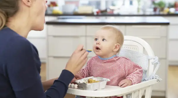 Mama karmi łyżeczką dziecko, które jeszcze nie siedzi. Maluch w kraciastym śliniaczku z zaciekawieniem patrzy na jedzenie.