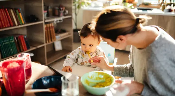 Mama karmi dziecko łyżeczką. Na stole są owoce i warzywa, idealne dla niemowlaka, który zaczyna poznawać, co może jeść 6 msc dziecko.