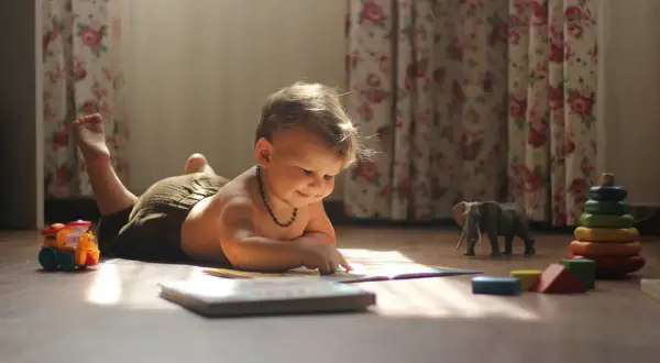 Małe dziecko leżące na brzuchu, podnoszące główkę, z zaciekawieniem przegląda książeczkę. Obok leżą zabawki: pociąg, słoń i wieża z kółek.