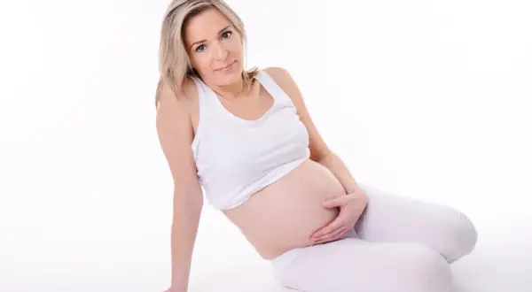 Kobieta w ciąży w białym topie i legginsach, siedząca na białym tle, trzymająca się za brzuch.