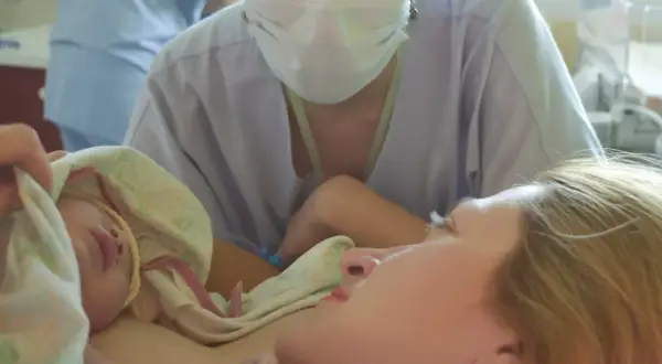 Noworodek przytulony do mamy, otoczony pieluchą. W tle widać personel medyczny w maseczkach, wspierający poród lotosowy.