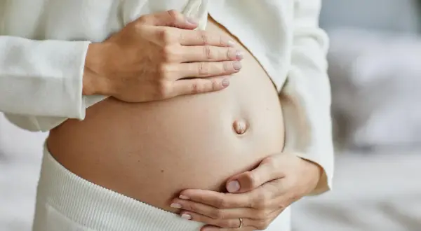 Zbliżenie na ciążowy brzuch kobiety, która delikatnie go obejmuje dłońmi.