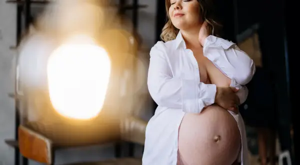 Kobieta w ciąży, z zamkniętymi oczami, czule obejmuje swój brzuch. Na skórze widoczne są drobne grudki, typowe dla **brodawek w ciąży**.