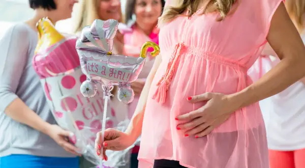 Ciężarna kobieta w różowej sukience trzyma balon w kształcie wózka z napisem "it's a girl!".