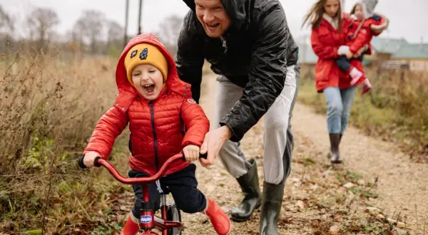 Radosne dziecko na rowerku biegowym, kiedy dziecko stoi bez podparcia, z tatą asekurującym. W tle mama z drugim dzieckiem.