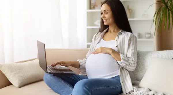 Kobieta w zaawansowanej ciąży sprawdza w internecie informacje o terminie porodu, siedząc na kanapie z laptopem na kolanach.