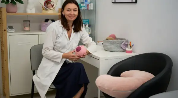 Położna podczas wizyty patronażowej pokazuje model piersi. W gabinecie widać pomoce dydaktyczne.