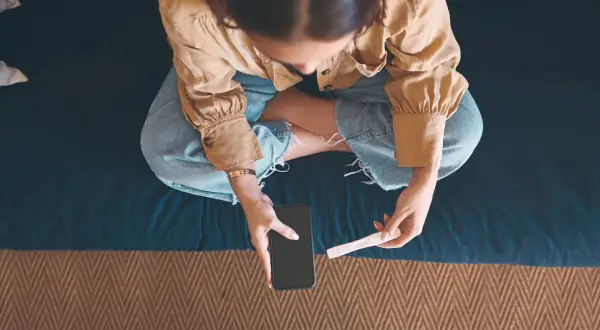 Kobieta siedzi ze skrzyżowanymi nogami, trzymając w dłoniach telefon i test ciążowy.