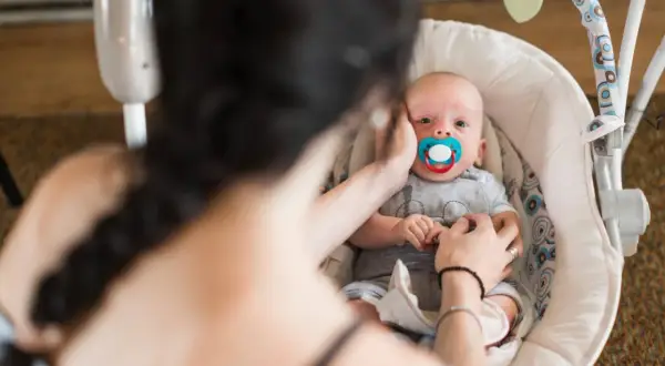Maluszek w bujaczku polecanym przez fizjoterapeutów, z niebieskim smoczkiem. Mama delikatnie głaszcze go po główce.