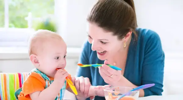 Mama karmi niemowlę, które próbuje samodzielnie jeść. To obrazek z tabeli żywienia 7-miesięcznego dziecka.