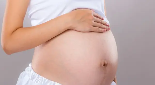 Brzuch kobiety w białej bluzce i spódnicy. Dłoń spoczywa na brzuchu, sugerując który tydzień ciąży.
