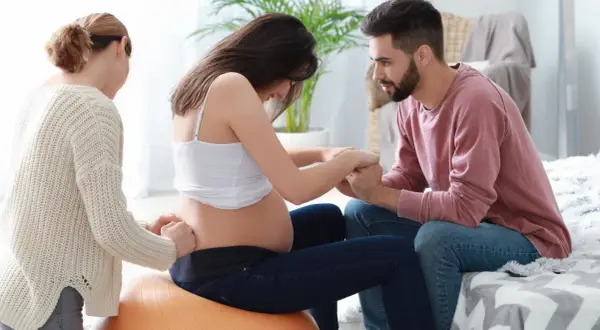 Kobieta w ciąży na piłce, czuć ruchy dziecka. Partner i doula wspierają ją.