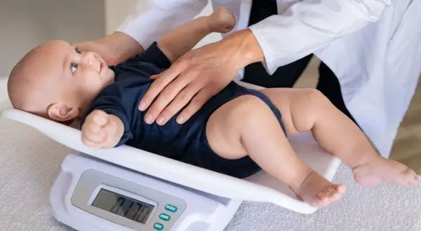 Lekarz waży niemowlę na wadze cyfrowej. Wynik 7.70 kg sugeruje, ile waży 10 miesięczne niemowlę.