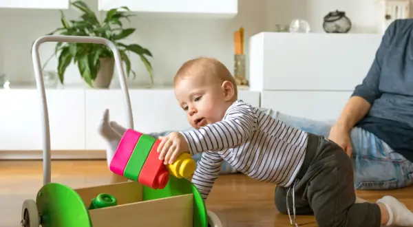 Radosne zabawy z 8 miesięcznym dzieckiem i jego nowym wózkiem-chodzikiem. Maluch z fascynacją wkłada kolorowe klocki do drewnianej skrzyni.