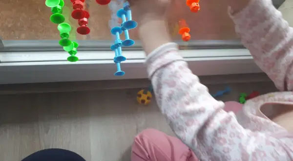 Rączki 15-miesięcznego dziecka bawią się kolorowymi przyssawkami na oknie. Radość i nauka przez zabawę.