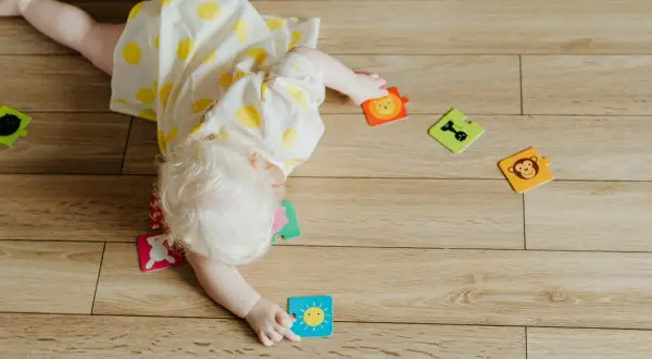 Niemowlę w żółtej sukience w kropki bawi się na podłodze, próbując obrócić się z brzucha na plecy. Wokół leżą kolorowe puzzle ze zwierzątkami.